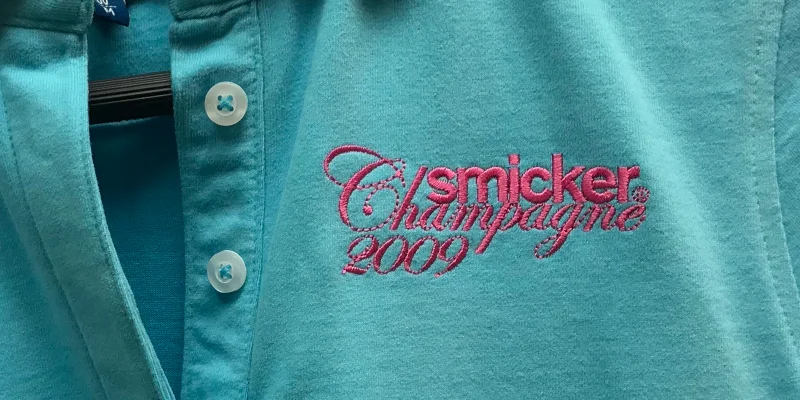 Rosa broderad logotyp på turkosblå micro textured pikétröja.