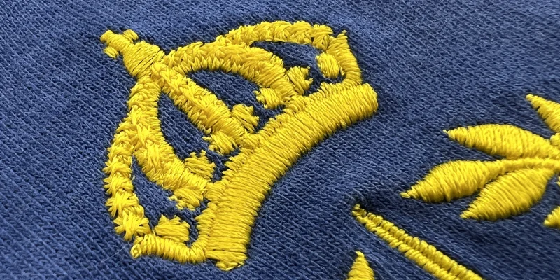 Närbild av broderad logotyp föreställande en gul kungakrona på blår piké.